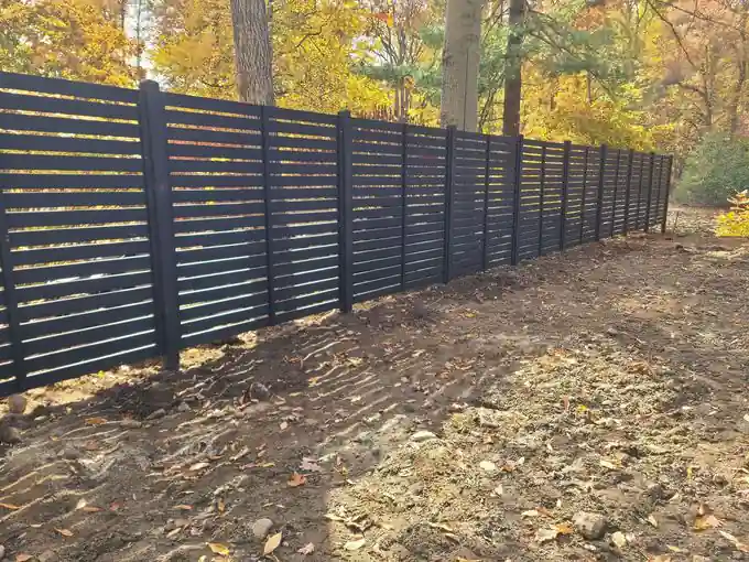 Long run of black horizontal-slat privacy fence on freshly graded soil under autumn trees