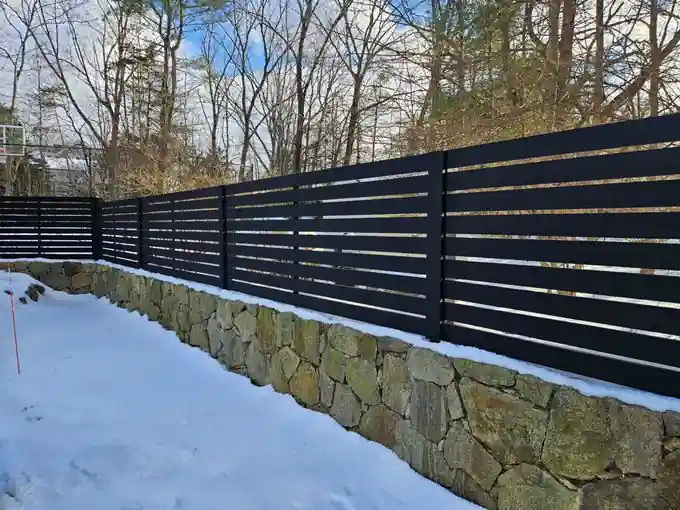 Black horizontal-slat privacy fence on a fieldstone retaining wall, snow on the ground