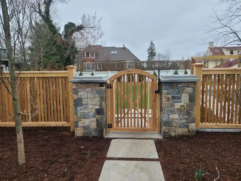 Arched cedar entry gate hung between two fieldstone piers, cedar picket fence on either side