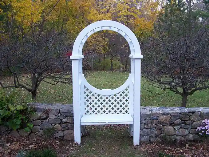 White lattice arbor with a round opening set on a fieldstone wall among autumn trees