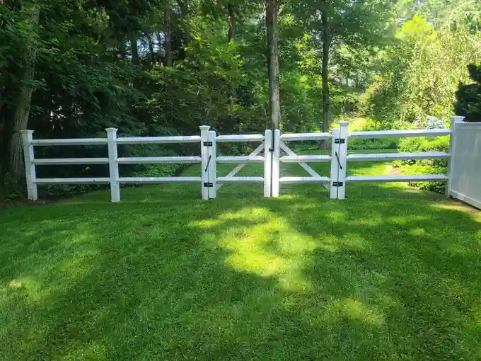 White vinyl three-rail ranch fence with a double gate across a sunlit lawn, woods behind
