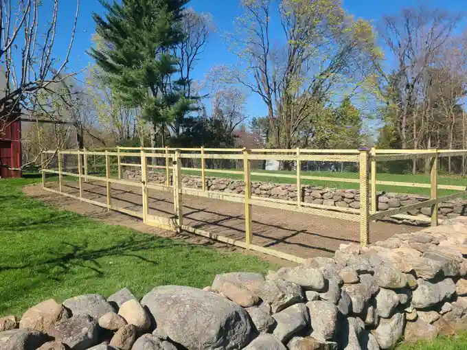 Tall natural-wood wire deer fence enclosure with a gate, built along an old fieldstone wall